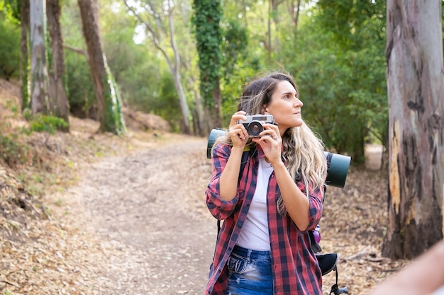 Free Photo pensive lady holding camera, looking around and standing on road. female tourist exploring nature and taking photo of forest. tourism, adventure and summer vacation concept