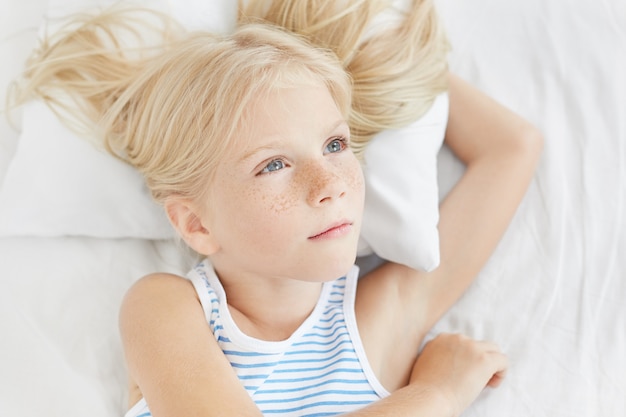 Pensive little girl with blue eyes and long eyelashes, having long blonde hair, wearing sailor T-shirt, lying on white pillow, looking aside