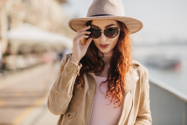 Pensive long-haired woman touching her sunglasses while walking on embankment