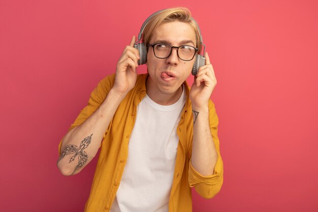 pensive looking at side young blonde guy wearing yellow t-shirt and glasses with headphones showing tongue isolated on pink with copy space