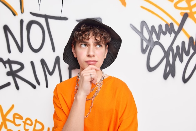 Free photo pensive man dressed in street style outfit holds chin wears black hat and orange tshirt poses next to graffiti wall has thoughtful expression