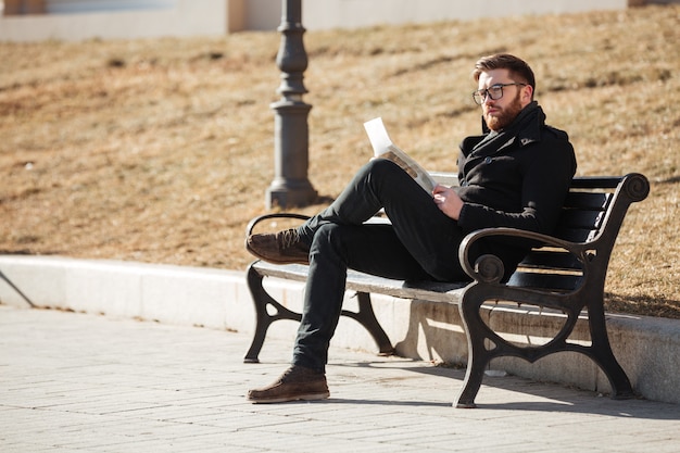 Pensive man sitting on bench and reading newspaper outdoors