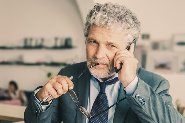 Pensive mature businessman talking on mobile phone, standing at co-working, leaning on desk