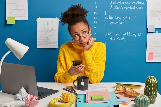 pensive office worker thinks on creative idea, uses mobile phone, waits for call, prepares for course work, learns languages online, spends time for autodidact drinks tea wears yellow sweater