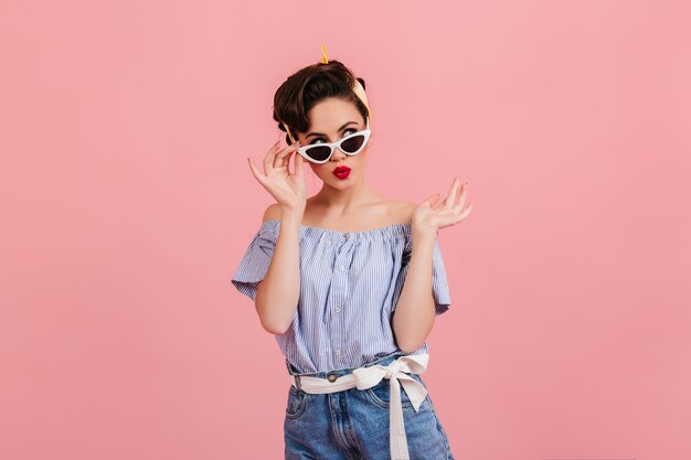 Pensive pinup girl in sunglasses standing on pink background. Studio shot of brunette young lady in summer outfit.