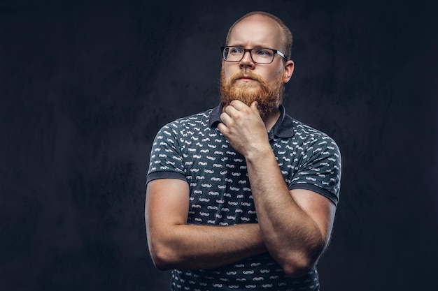 Free Photo pensive redhead bearded male in glasses dressed in a t-shirt posing with a hand on chin. isolated on a dark textured background.