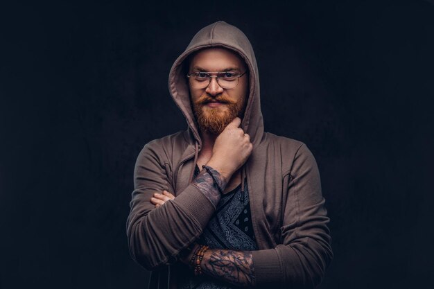 Pensive redhead hipster with full beard and glasses dressed in hoodie and t-shirt poses with hand on chin in a studio. Isolated on a dark background.