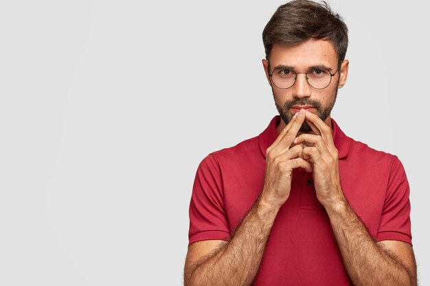 Pensive serious bearded male keeps hands near mouth, contemplates about something, dressed in red t-shirt, stands against white wall