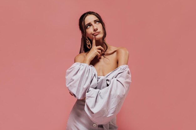 Pensive slim lady with tanned skin and brunette hair in gold accessories in wide sleeve lilac shirt looking up and posing