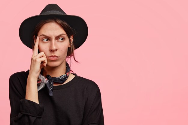 Pensive western woman keeps fore finger on temples, contemplates about something with thoughtful expression, has serious look, wears clothes