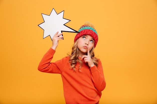 Pensive Young girl in sweater and hat holding blank speech star and looking up over orange