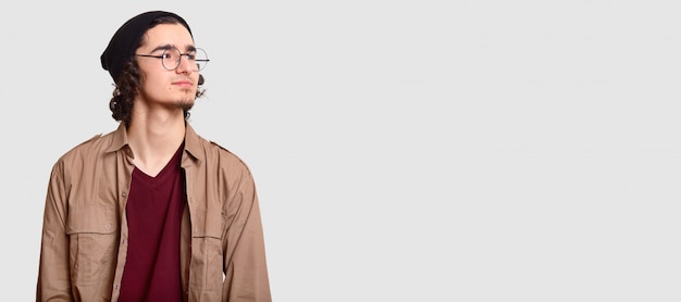 pensive young hipster looks pensively aside, wears round spectacles, casual clothes, isolated on white with free space for your advertising content or promotion