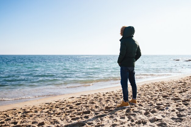 Pensive young man looking at the sea, standing on the sand. Back view.