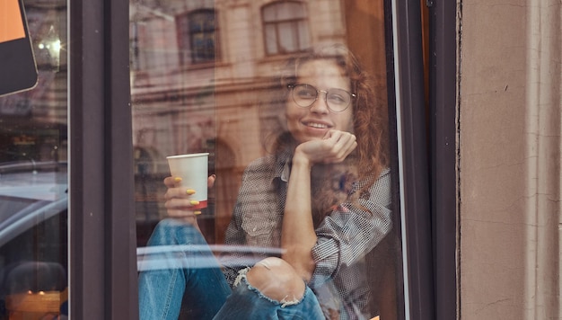 Free photo pensive young redhead curly girl wearing casual clothes and glasses sitting on a window sill with hand on chin, holds a takeaway coffee.