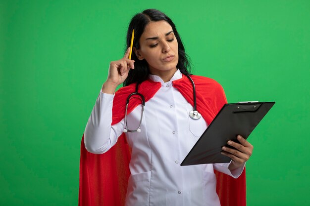 pensive young superhero girl wearing medical robe with stethoscope holding and looking at clipboard putting pen on head isolated on green