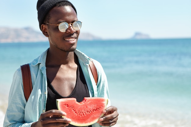 People, active modern lifestyle, travel, vacations and tourism concept. Cheerful young dark-skinned backpacker in stylish clothing spending sunny summer day at seaside, enjoying juicy watermelon