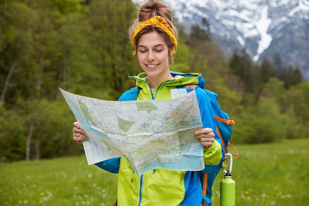 Free photo people, adventure and trekking concept. happy female tourist holds paper map, strolls on valley near mountains