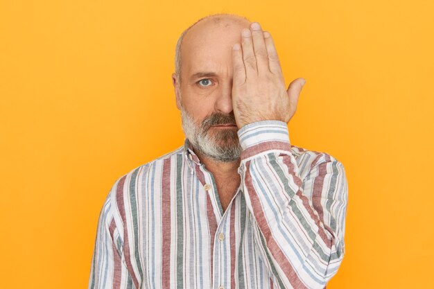 People, age, health and retirement concept. Unshaven bald retired man in striped shirt covering one eye with hand having vision tested in ophthalmology clinic, not able to see nearby objects