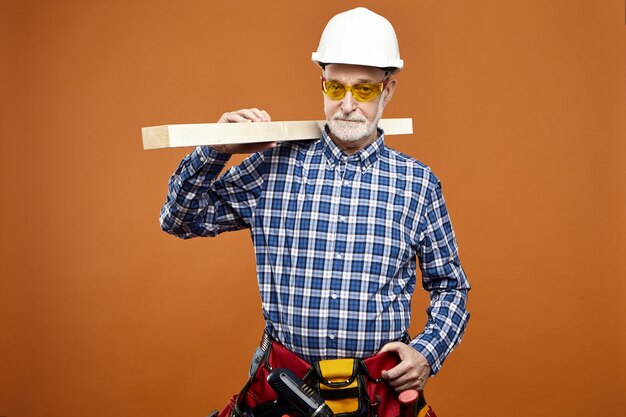 People, age, job and occupation concept. Portrait of confident focused senior man wearing yellow goggles, plaid shirt, hardhat and wiast bag with tools, carrying wooden bar on his shoulder