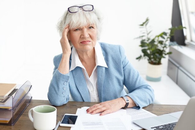 People, age, job, stress and health concept. Picture of displeased gray haired businesswoman frowning, touching head to ease pain because of headache, working too much, studying papers in office