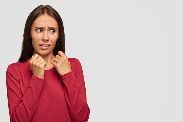 People and aversion concept. Displeased attractive female looks with scared negative facial expression, dressed in red sweater, stands against white wall