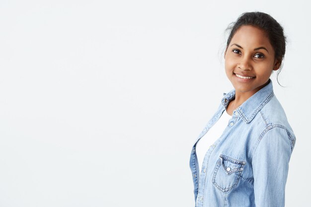 People and beauty concept. Portrait of beautiful dark-skinned woman posing on white wall  with cute and charming smile, dressed in light-blue denim shirt.