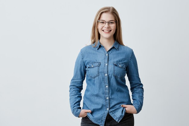 People, beauty and lifestyle concept. Attractive sensual blonde woman with spectacles and wide smile dressed in denim shirt smiling broadly being happy to meet her best friend. Joyful nice female