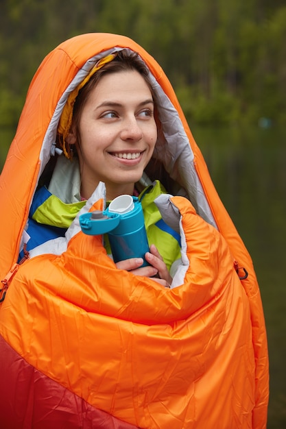 Free photo people and camping concept. glad lovely female hiker wrapped in orange sleeping bag, warms herself during cold day