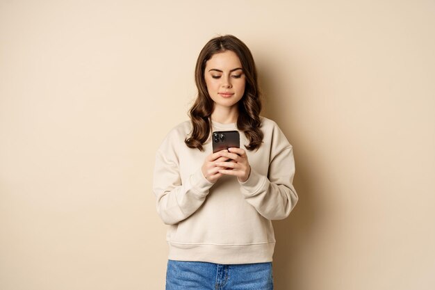 People and cellular technology. Beautiful stylish woman using mobile phone, smartphone app, smiling and looking at screen, standing over beige background