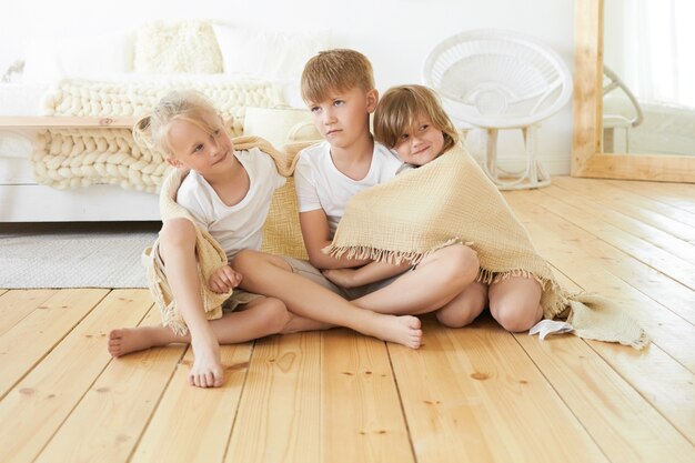 People, childhood, family, love and togetherness concept. Sweet cozy picture of three cute little children siblings sitting on wooden floor together wrapped in blanket, embracing each other