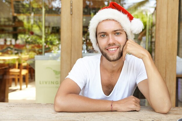 People, Christmas, New Year and holidays concept. Headshot of attractive young hipster with stylish beard wearing Santa Claus hat smiling cheerfully