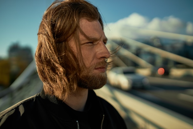 Free photo people and city lifestyle concept. portrait of attractive stylish bearded guy with messy red hair and nose piercing posign against urban landscape background, having serious confident look