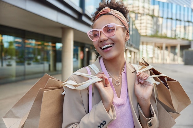 Free photo people consumerism and purchasing concept glad fashionable woman wears pink sunglasses stylish jacket carries paper shopping bags has happy mood strolls at sreeet against urban bbackground