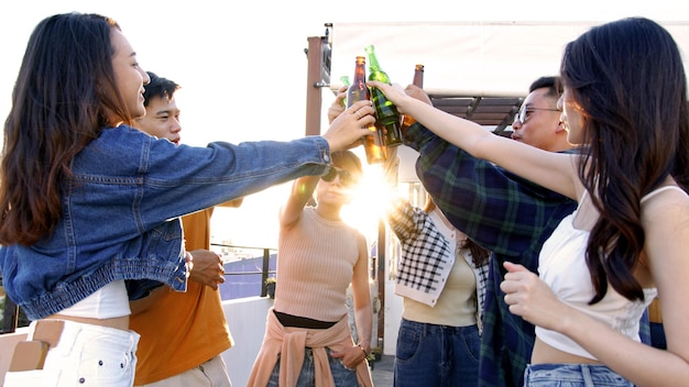 people drinking alcohol at sunset rooftop party asian friends clinking with beer bottles on terrace