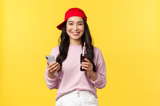 People emotions, drinks and summer leisure concept. Young teenage korean girl in red cap, messaging, using smartphone and drinking soda beverage, standing yellow background pleased.