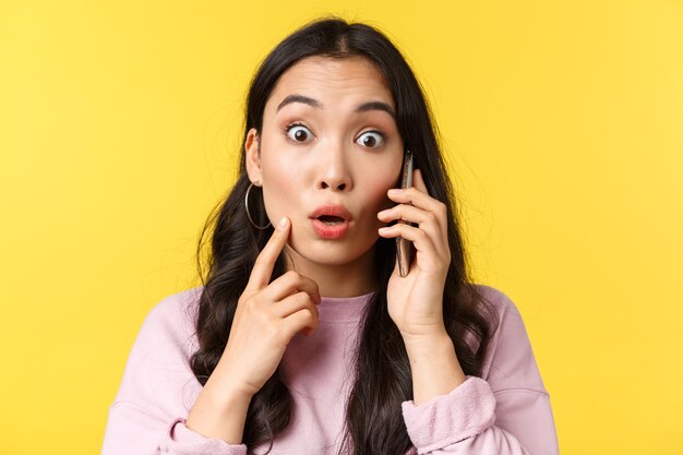 People emotions, lifestyle leisure and beauty concept. Close-up of shocked and surprised asian girl talking on phone, stare camera with popped eyes as hear big news, yellow background