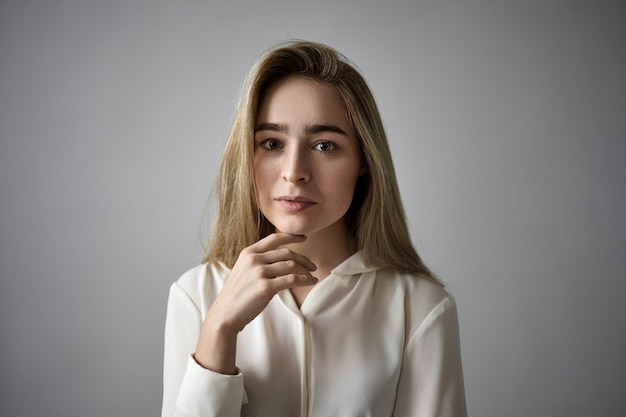 People, femininity, beauty, style and fashion concept. Horizontal shot of attractive fair haired young Caucasian woman with loose hair and freckles tocuhing her chin, having serious thoughtful look