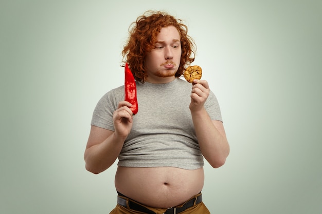 People, food, diet, nutrition and unhealthy lifestyle. sad indecisive young plump man with big stomach hanging out of jeans, facing difficult dilemma between vegetable and cookie