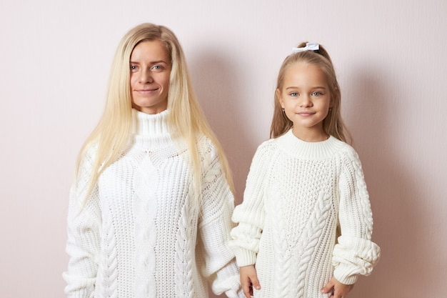 Free photo people and generations concept. isolated shot of attractive young european mother posing  holding hands with beautiful little daughter, both dressed in cozy warm sweaters