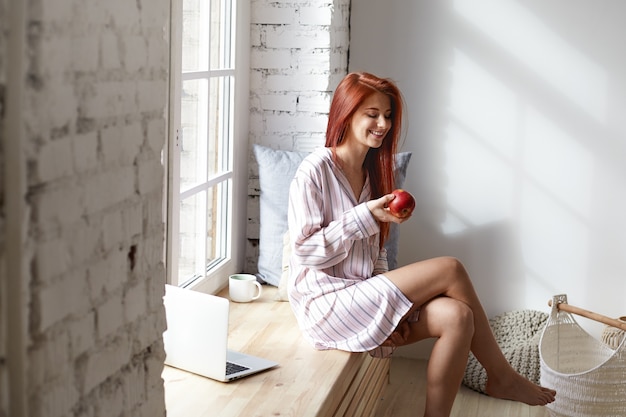 Free photo people, healthy lifestyle, food, diet and morning routine concept. beautiful red haired young caucasian woman in striped night suit laughing, enjoying calm peaceful morning at home, eating fresh apple