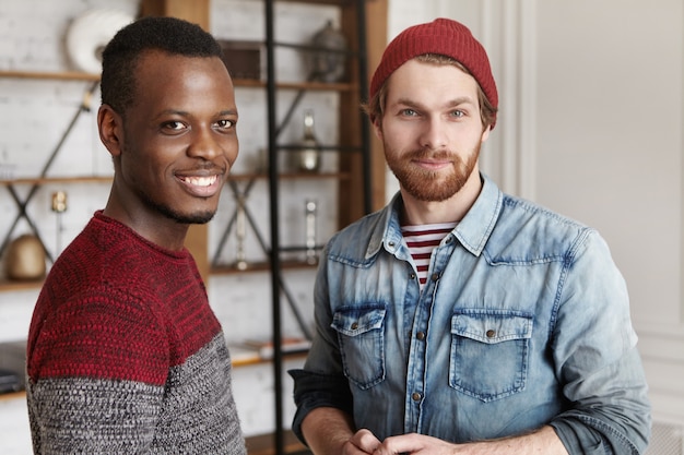 People and interracial friendship concept. Two old male friends that came across each other at cafe standing in modern cafe interior and having conversation, both looking with happy smiles