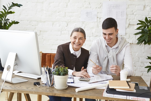 People, job, teamwork and cooperation concept. Happy middle aged female chief engineer having fun in office while explaining something to her young male assistant, sitting at desk with pc and papers