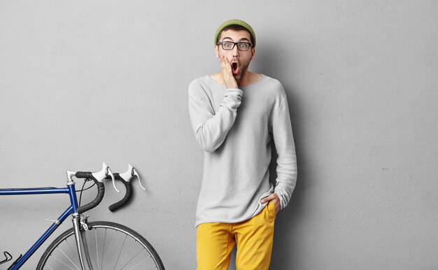 People, leisure, activity, sport concept. Young handsome man with beard, keeping one hand on cheek and other in pocket of yellow trousers, being shocked because of bad weather and spoiled weekends