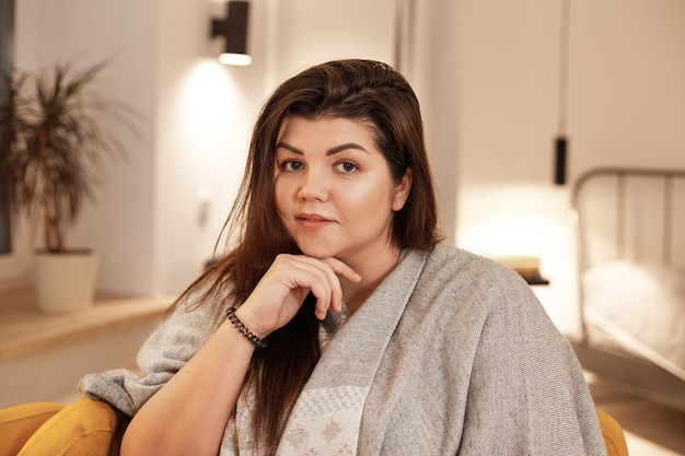 People, leisure, coziness and lifestyle concept. Indoor image of attractive cute young plus size female with loose black hair relaxing at home, sitting in armchair, wrapped in warm blanket