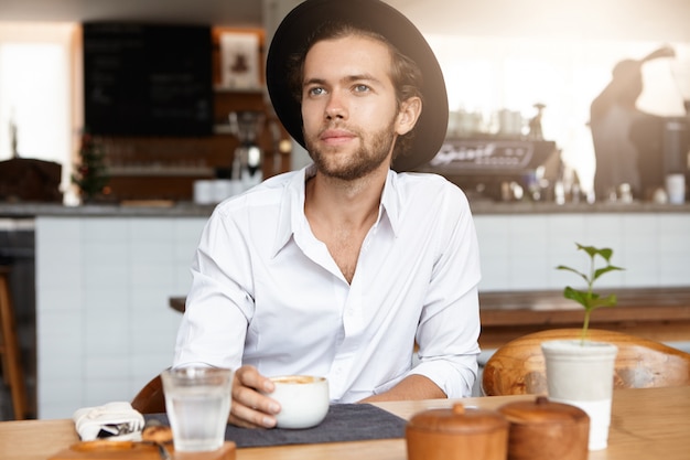 People, leisure and lifestyle concept. Indoor shot of handsome young bearded male wearing trendy hat sitting at wooden table with mug, enjoying fresh cappuccino during coffee break, having happy look