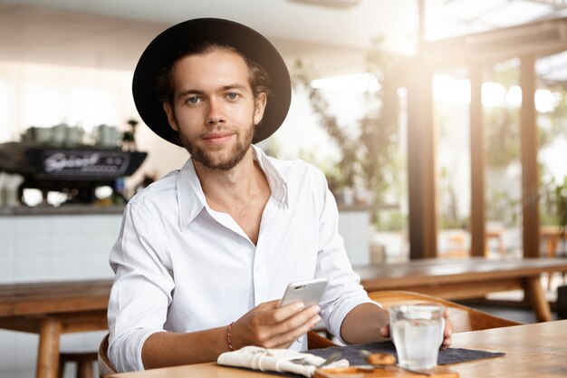 People, leisure and modern technology. Young student with happy look enjoying high speed internet connection on his smart phone. Fashionable man in trendy headwear using electronic device at cafe