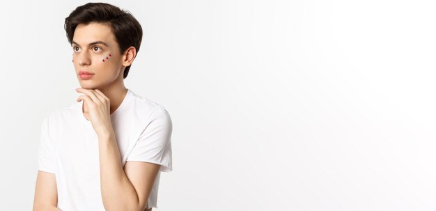 People lgbtq and beauty concept Beautiful androgynous man with glitter under eyes looking at upper left corner posing against white background