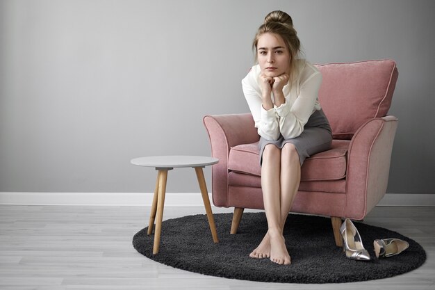 People and lifestyle concept. Beautiful young female teacher with hair bun relaxing at home after lectures at college, sitting barefooted in armchair, looking at camera with tired facial expression