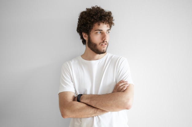 People and lifestyle concept. Portrait of fashionable young Caucasian bearded hipster wearing white t-shirt posing with arms folded, looking away with serious thoughtful expression