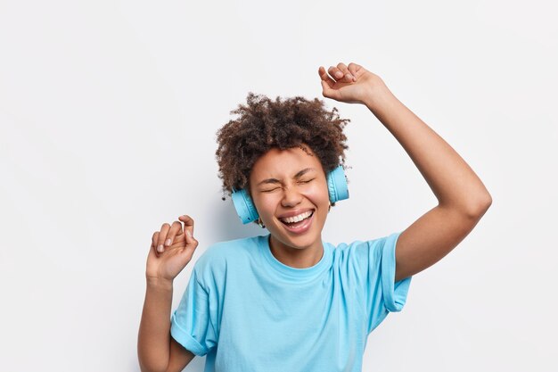 People lifestyle leisure concept. Energetic cheerful African American woman with curly hair dances carefree raises arms feels amused enjoys favorite music wears wireless headphones blue t shirt